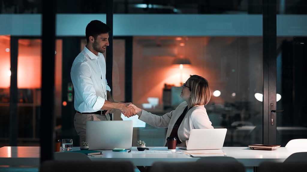 An individual sitting at their desk with a laptop, shaking hands with another professional individual standing slightly above them