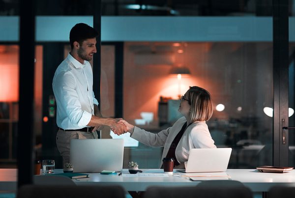 An individual sitting at their desk with a laptop, shaking hands with another professional individual standing slightly above them