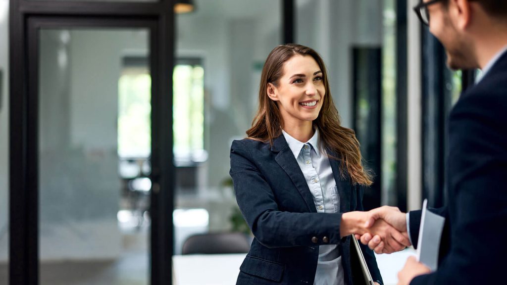 Professional, brunette woman in a modern office setting enthusiastically shakes hands with a professional gentleman