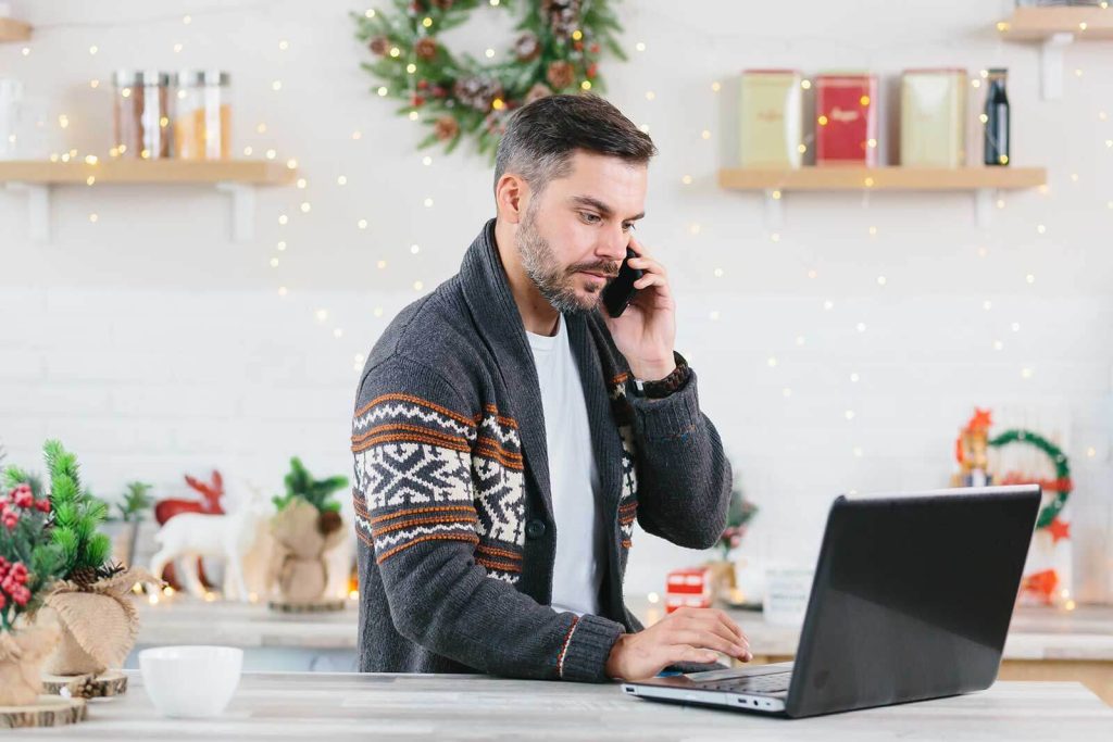 Man in holiday attire sits at kitchen counter looking at his laptop while on the phone. Festive and holiday decor is in the background