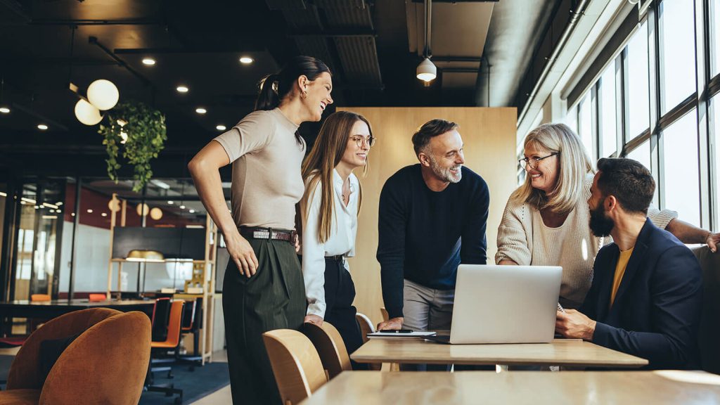 A mix of five professionals speaks pleasantly at a boardroom table with a laptop in front of them