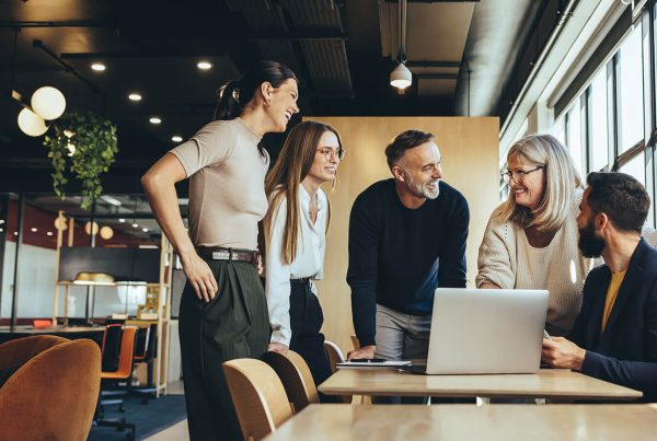 A mix of five professionals speaks pleasantly at a boardroom table with a laptop in front of them