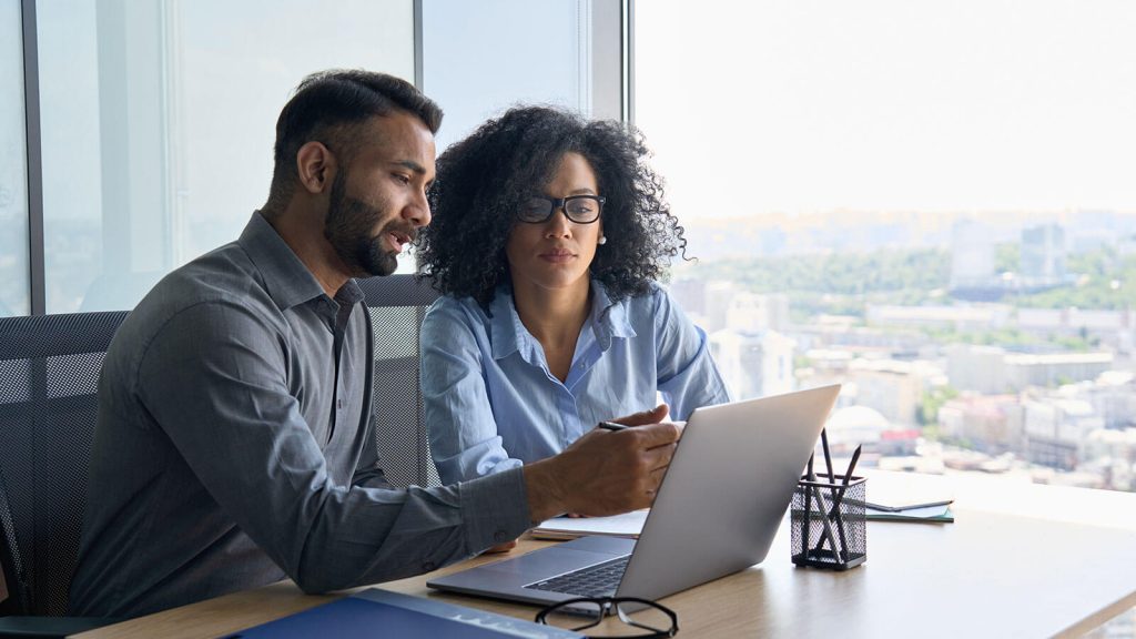 A male and female examine a document on their laptop in a professional office setting